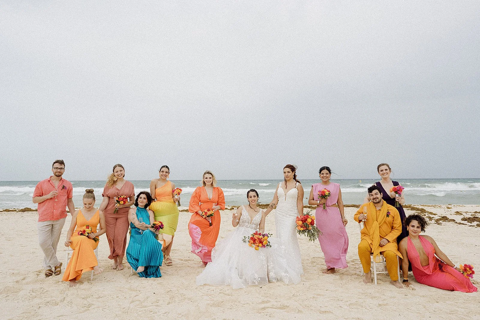 Grupo de personas en la playa en un día nublado, con dos mujeres en el centro con vestidos blancos de novia, rodeadas de amigos en ropa de colores brillantes, algunos con flores en las manos, todos sonriendo y posando para la foto.