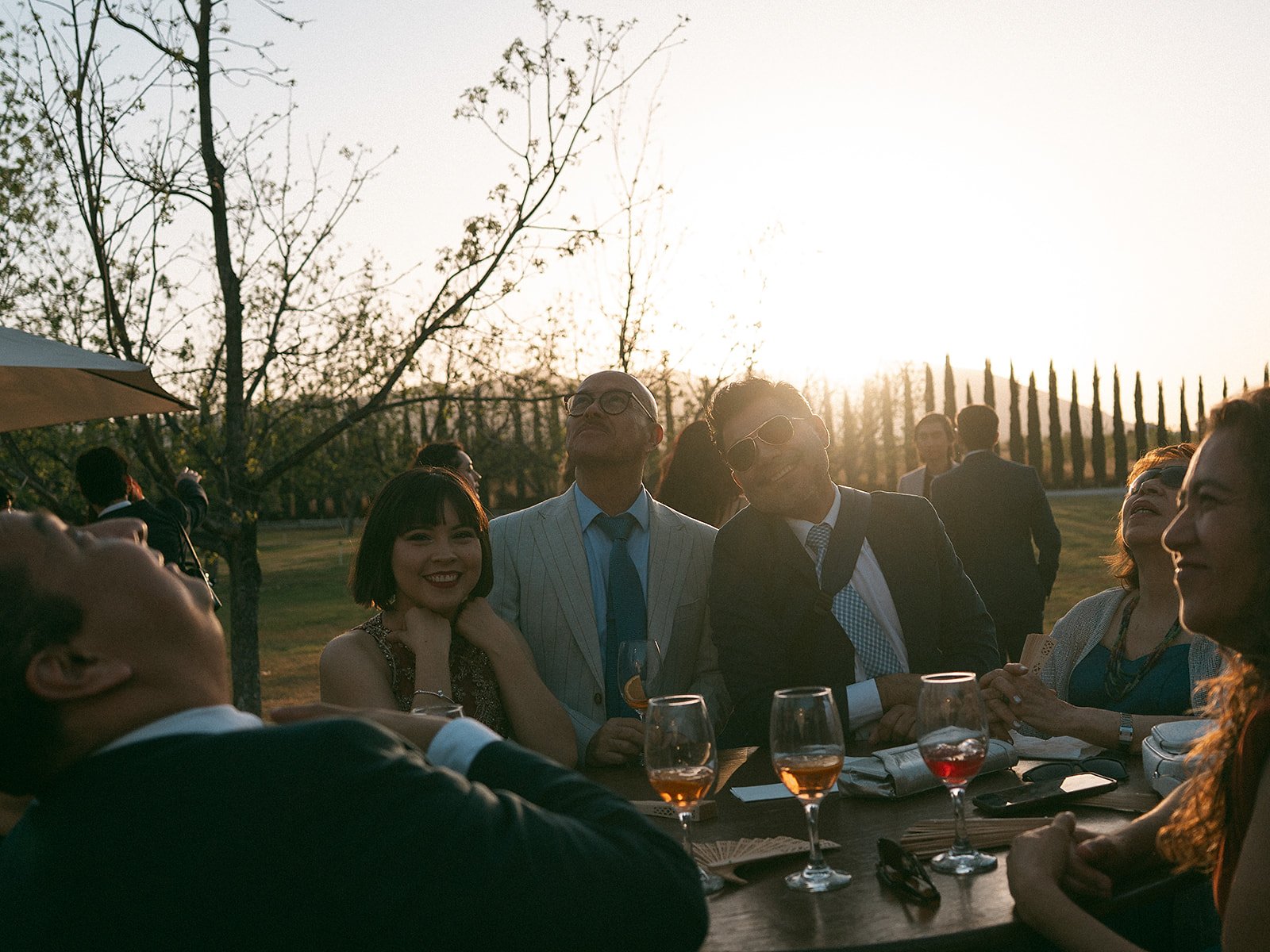 Grupo de personas en una reunión social al aire libre en la tarde, con varios vasos de vino y conversación animada.