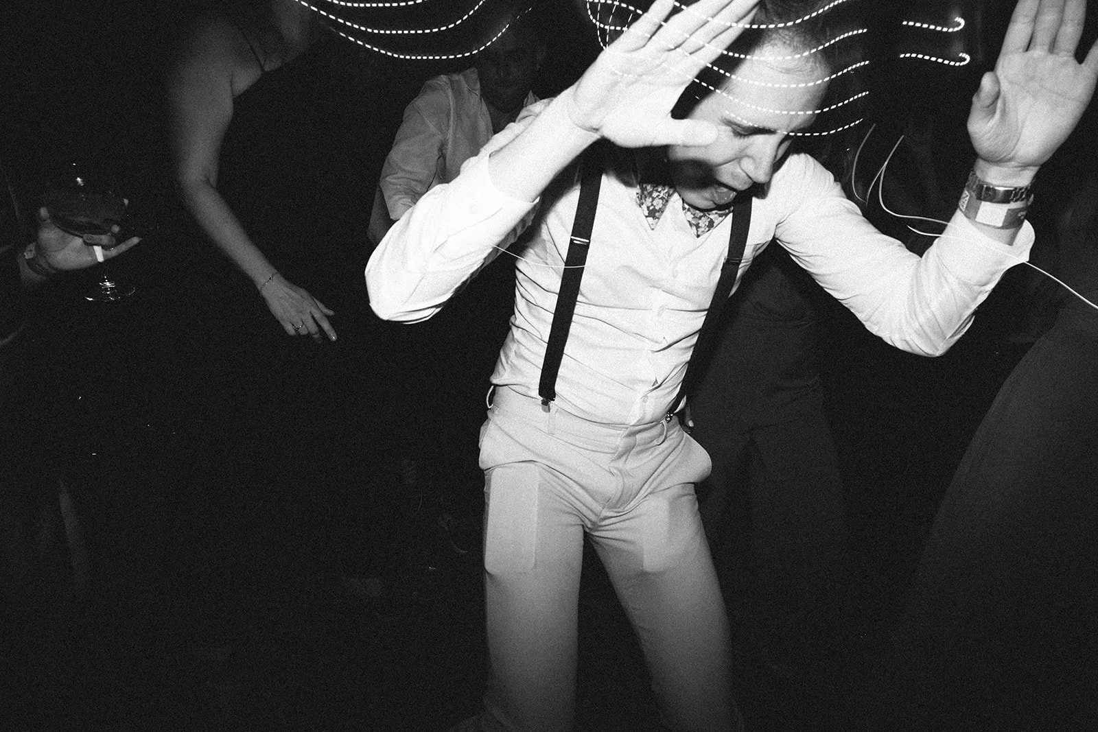 Hombre bailando en una fiesta, vestido con camisa blanca, pantalones claros y tirantes, rodeado de otros asistentes.