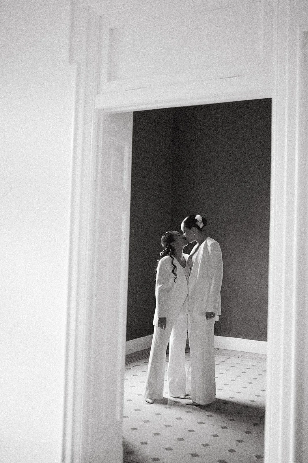 Una pareja con ropa formal, una mujer y una mujer, se besan en una habitación iluminada, vista a través de una ventana o espejo.
