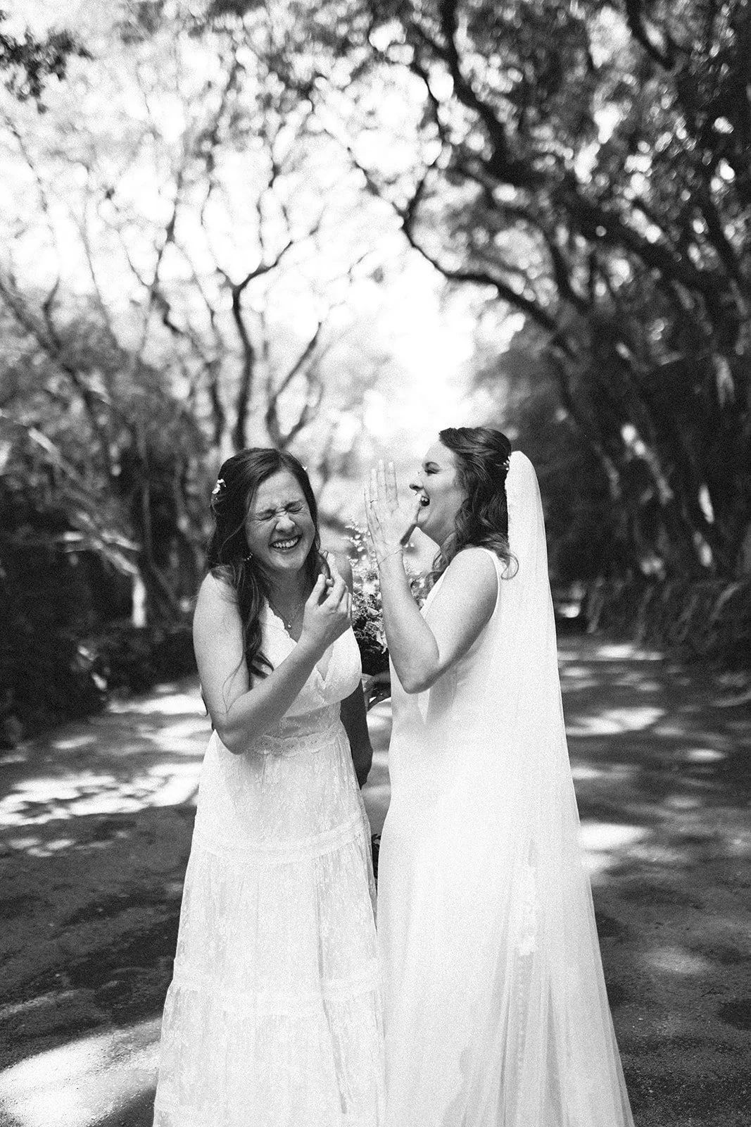 Dos mujeres vestidas con vestidos de novia riendo y compartiendo un momento alegre en un sendero rodeado de árboles, en una fotografía en blanco y negro.