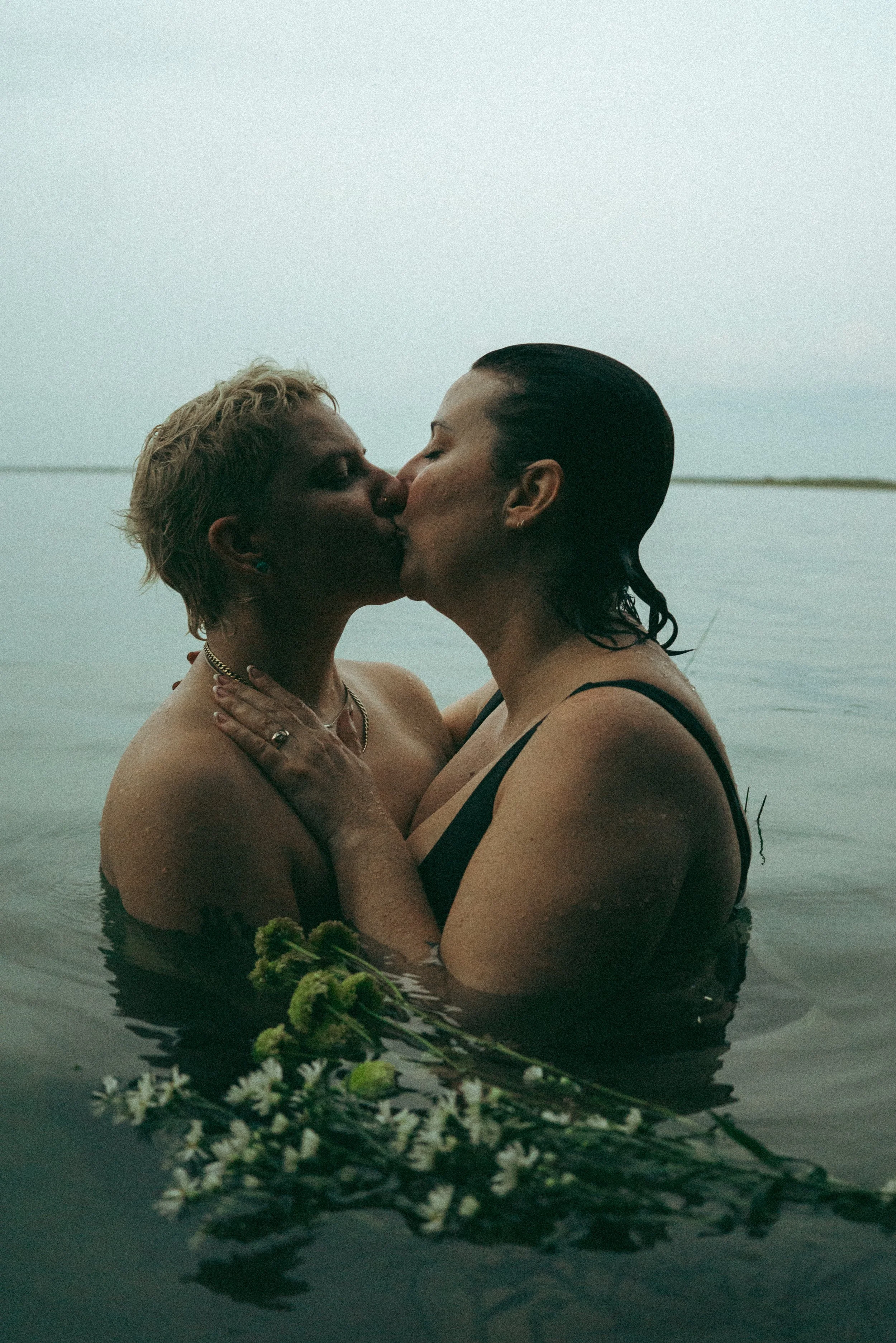 Dos mujeres en el agua, compartiendo un beso, con un fondo de cielo gris y agua tranquila, rodeadas de flores.
