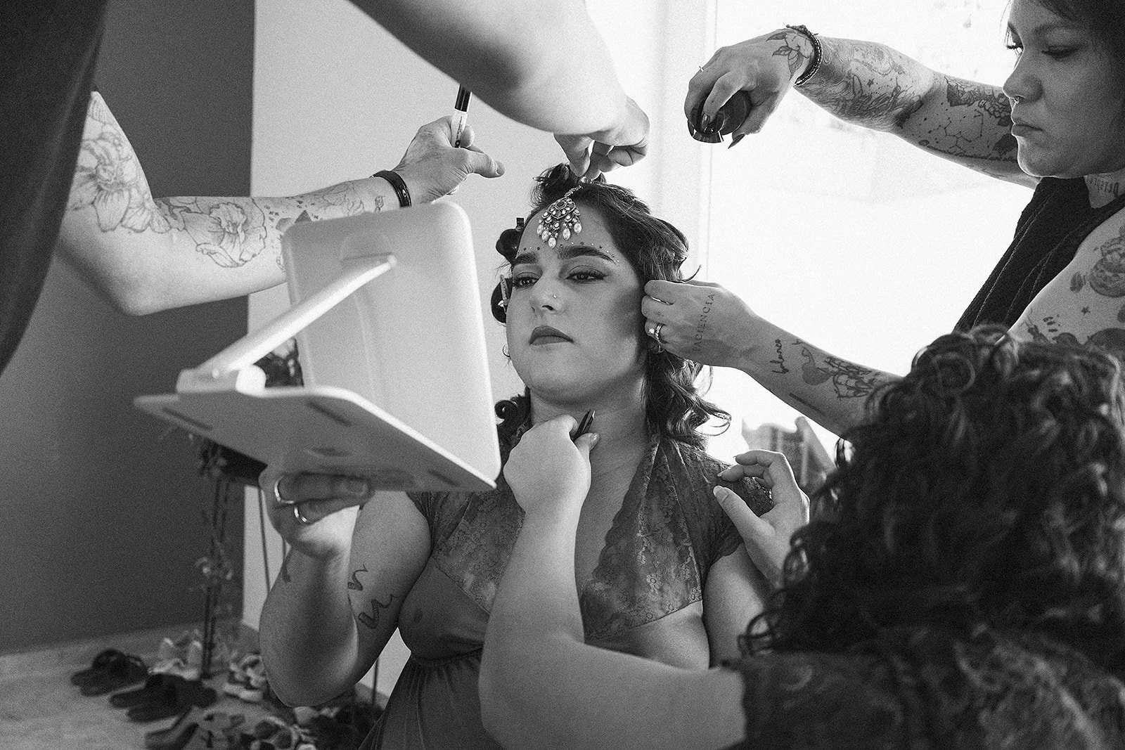 Mujer en sesión de maquillaje y peinado, con varias personas ayudándola, usando un tocado y vestido, en un entorno de preparación de evento.