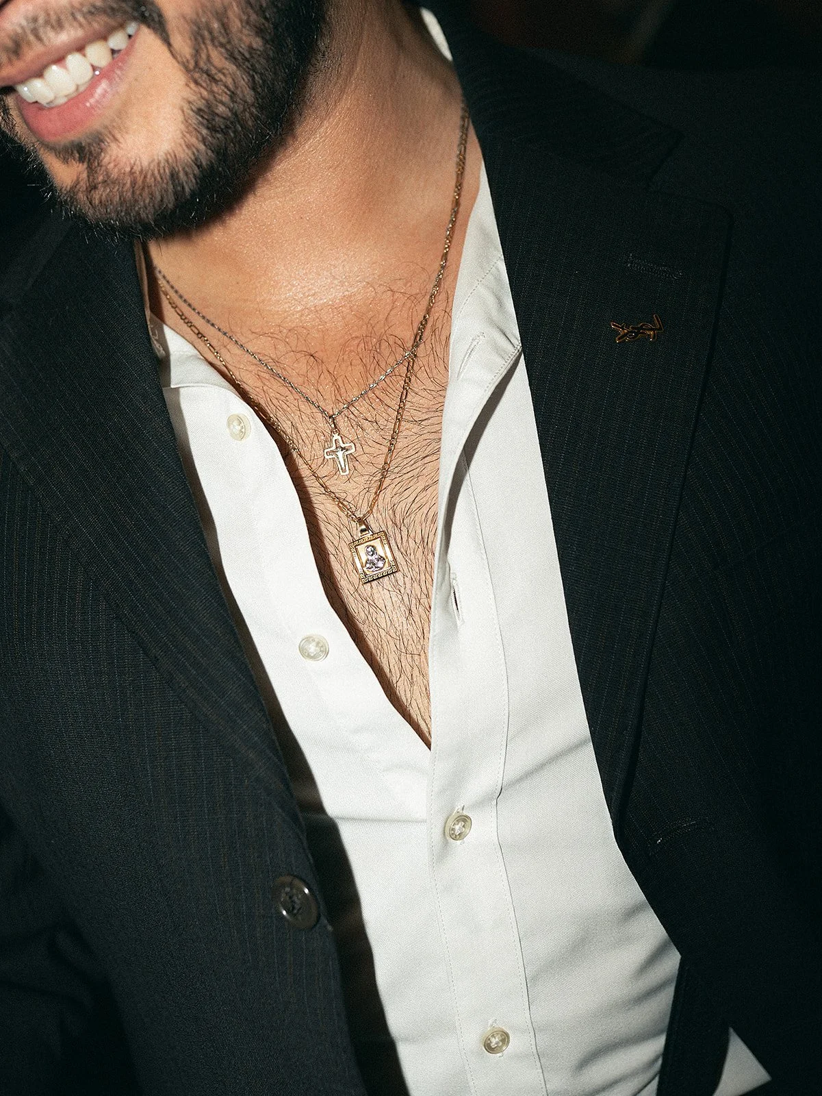 Hombre con barba que viste saco negro, camisa blanca y lleva varias cadenas de oro con cruces y colgantes.