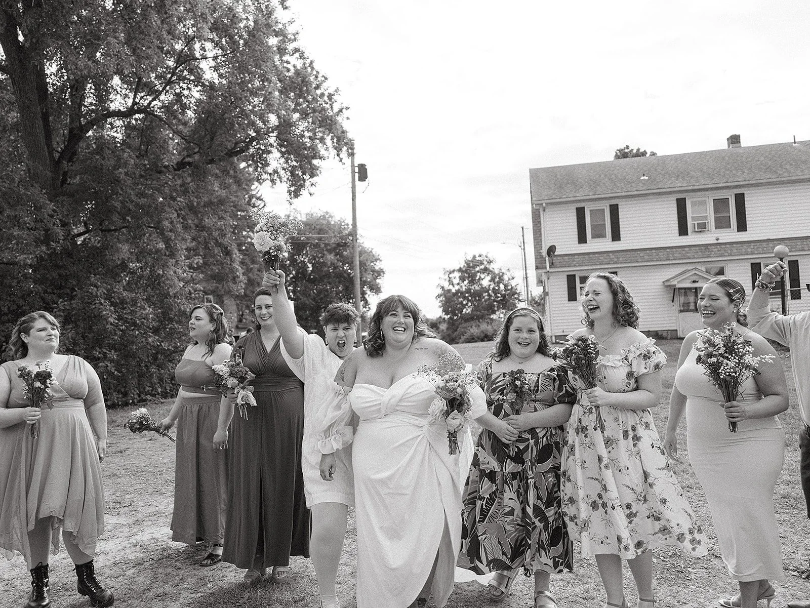 Un grupo de mujeres, aparentemente en una boda, caminando juntas, sonriendo y sosteniendo ramos de flores en un entorno exterior con árboles y una casa en el fondo.
