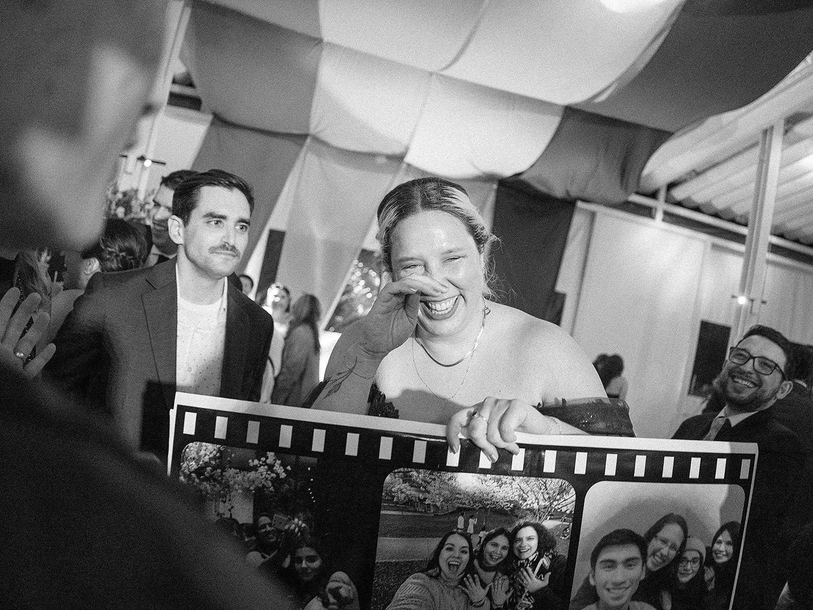 Una mujer riendo y cubriéndose la cara con la mano en un evento social, rodeada de varias personas que también sonríen.