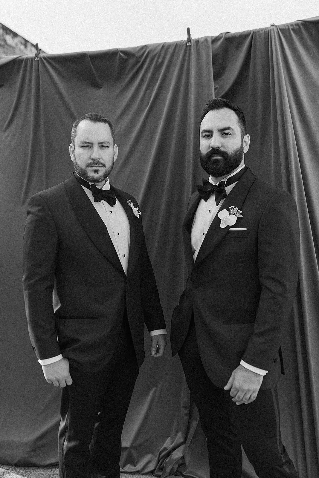 Dos hombres con trajes formales y pajaritas, en un evento especial, posando frente a una cortina.