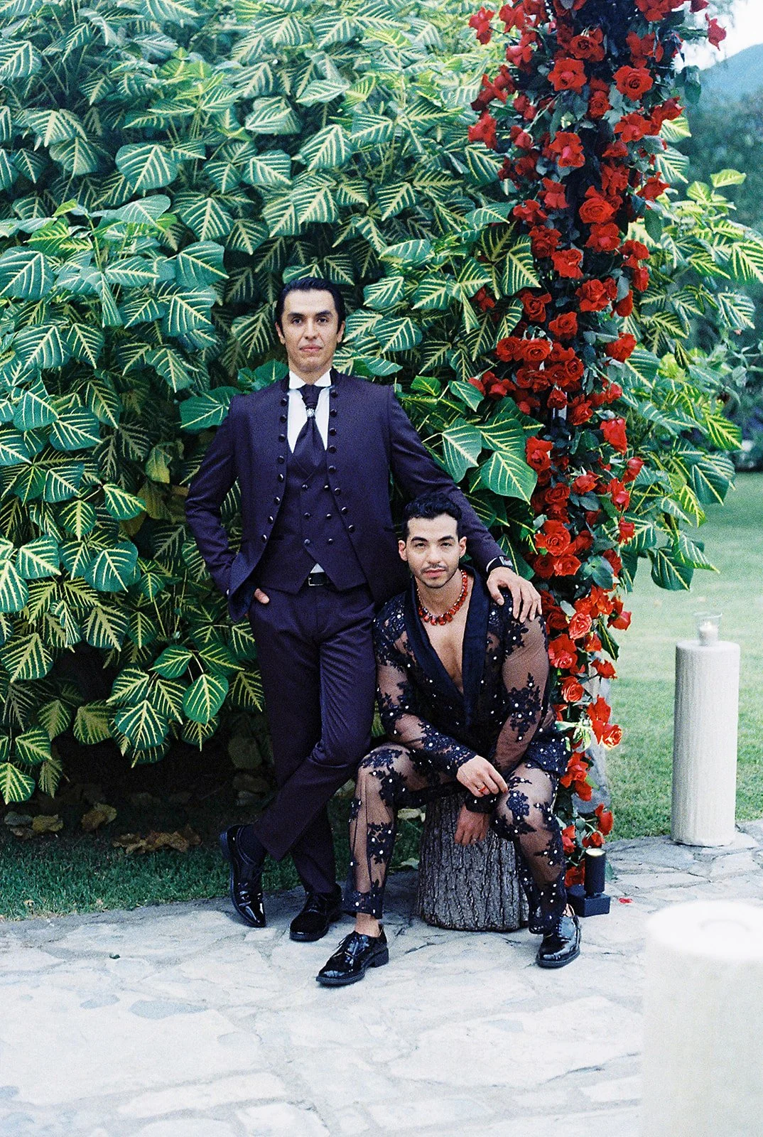 Dos personas posando en un jardín con arbustos verdes y flores rojas. Una persona está de pie vestida con un traje azul oscuro y la otra está sentada en un tronco con un vestido transparente decorado con flores negras y un collar rojo.