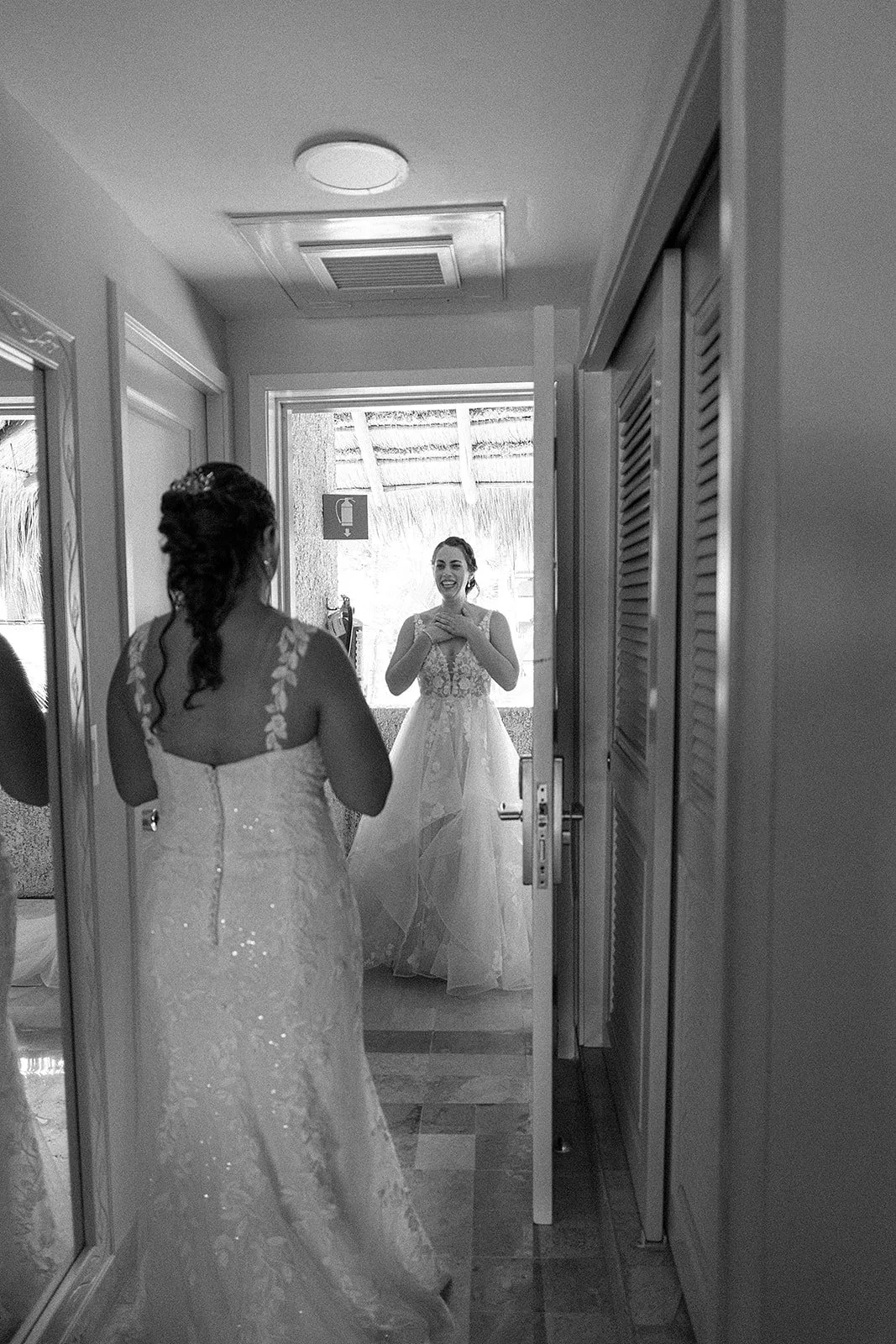 Dos mujeres en un vestidor, una de ellas viste un vestido de novia y la otra, también con vestido de novia, se mira en el espejo con una expresión de felicidad.