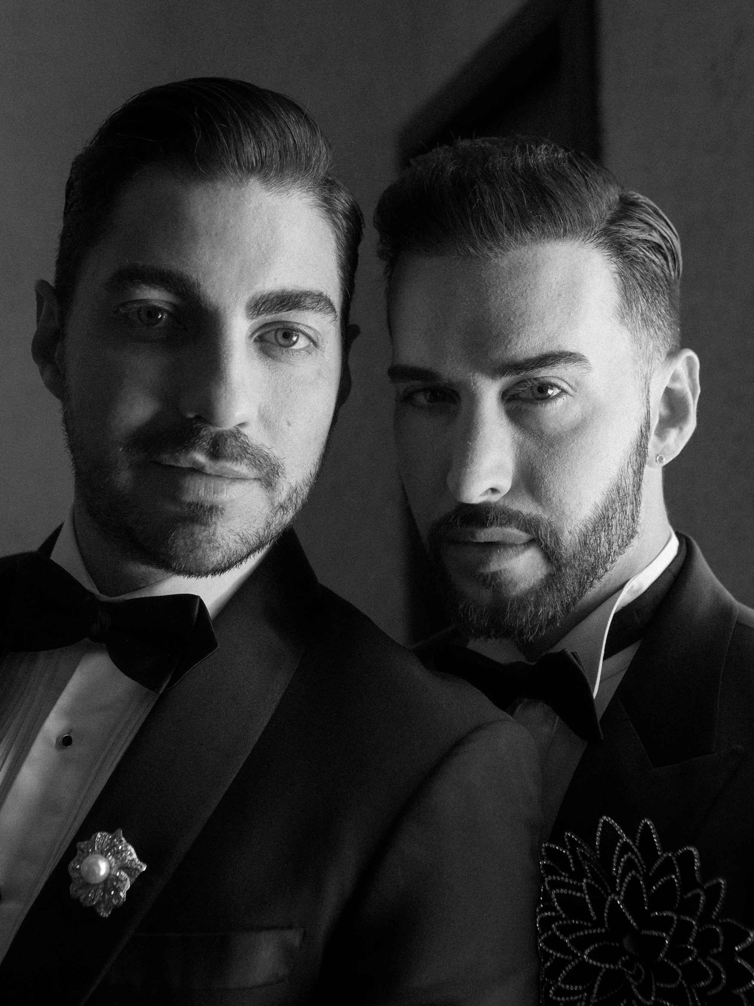 Retrato en blanco y negro de dos hombres con barba y cabello peinado, vestidos con tuxedos y corbatas, posando juntos para la foto, con fondo oscuro.