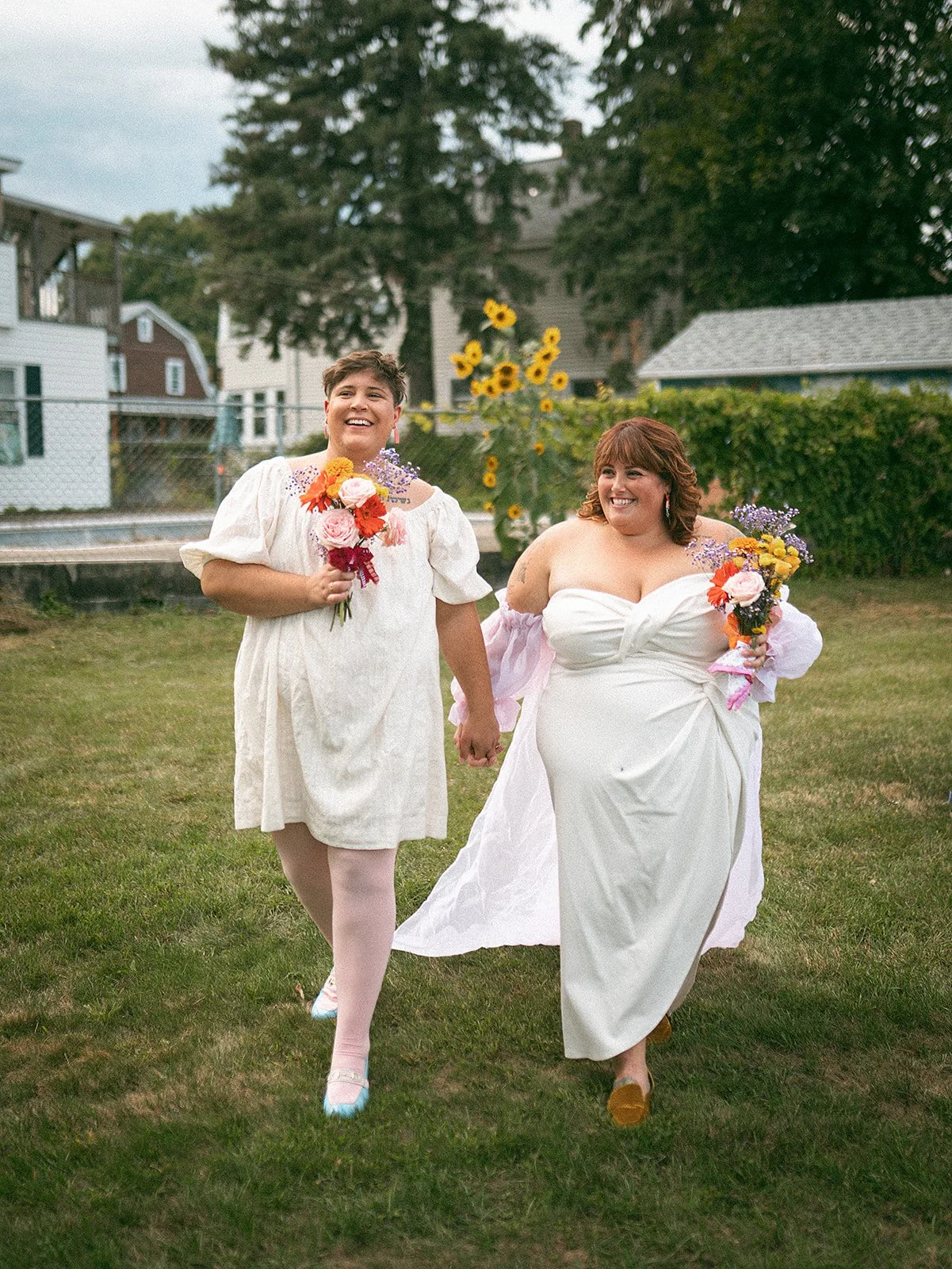 Dos mujeres con trajes blancos y flores en las manos pasean en un parque, una de ellas con un vestido de novia y la otra con un vestido suelto y color crema, ambas son felices.