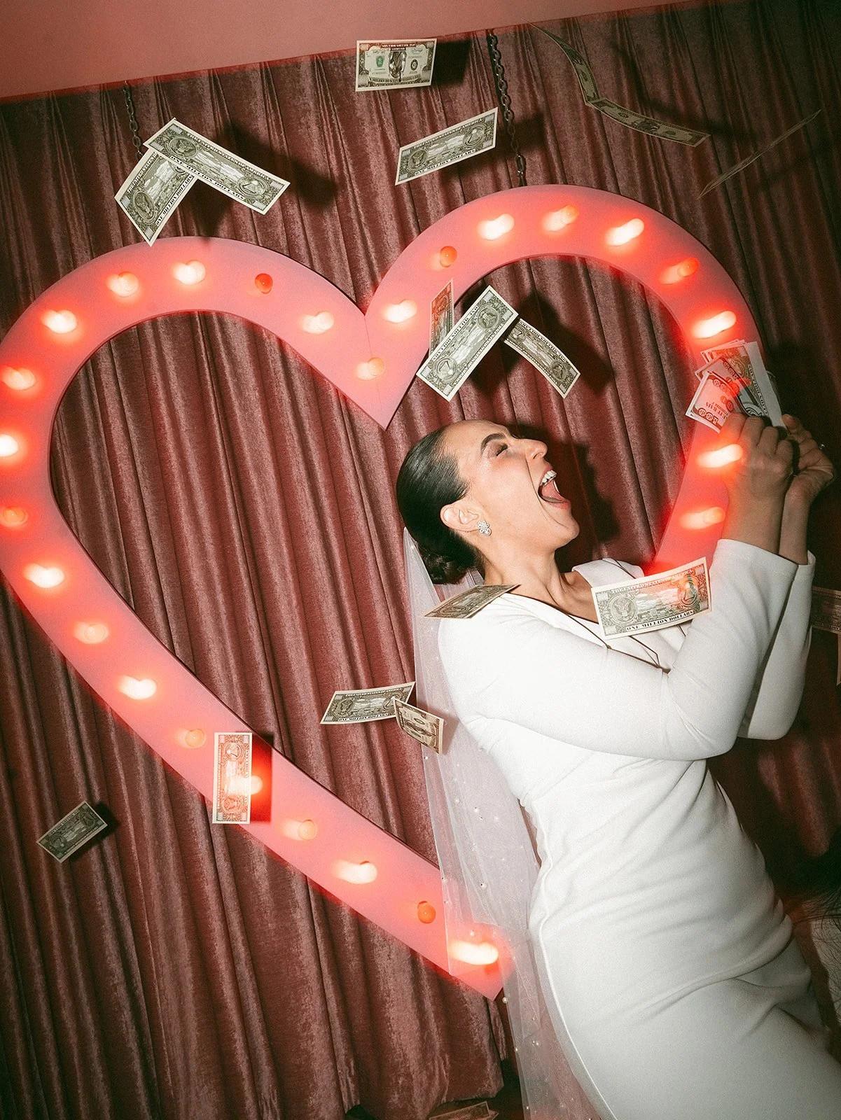 Mujer feliz celebrando, sosteniendo billetes de dólar, rodeada de una gran figura con luces en forma de corazón, con cortinas rojas de fondo.
