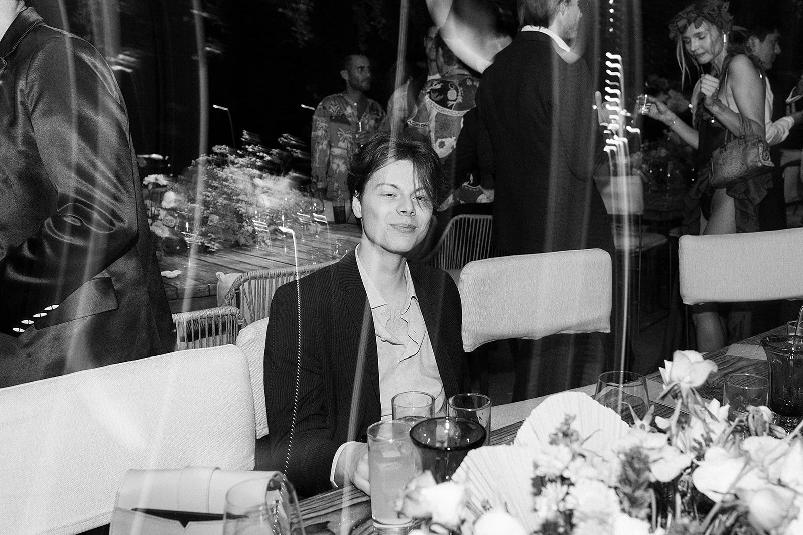 Mujer con ropa formal sentada en una mesa en una cena o evento social, con muchas personas en el fondo, en un ambiente elegante y decorado con flores.