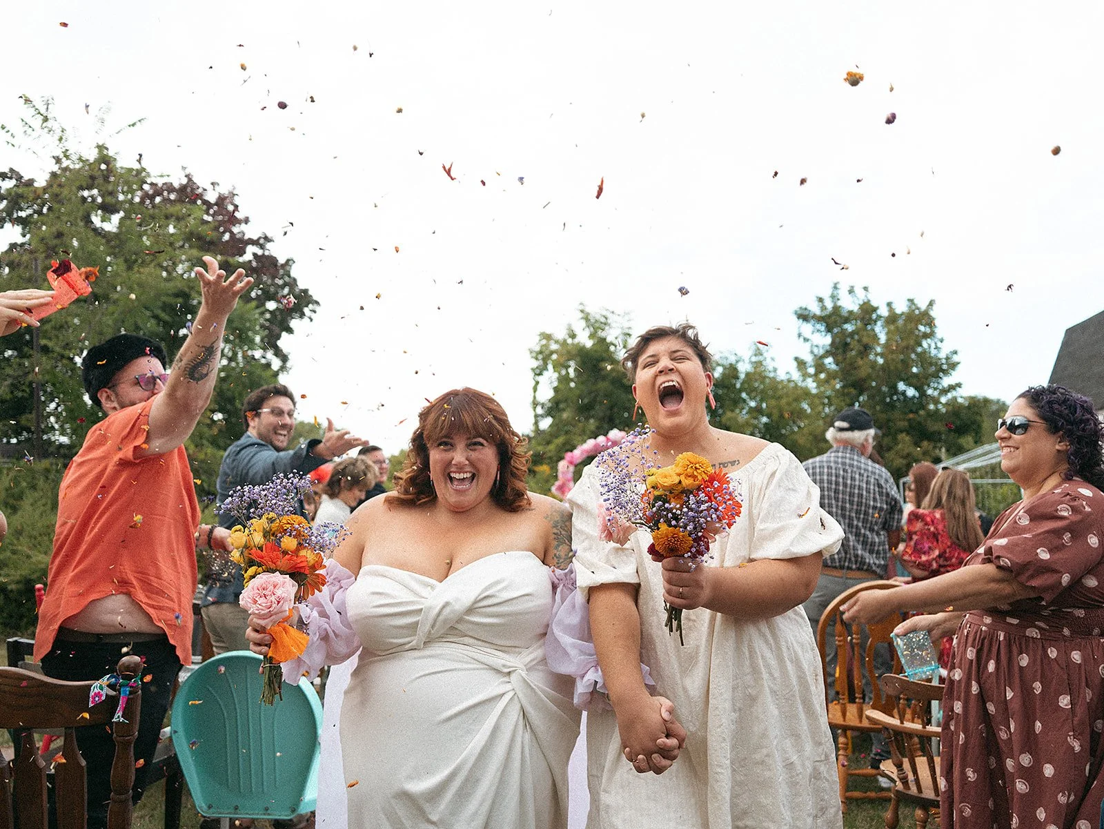 Pareja de mujeres en una boda, una con vestido blanco y la otra con vestido beige, tomadas de la mano y sonriendo, rodeadas de fiesta y confeti en un espacio al aire libre.