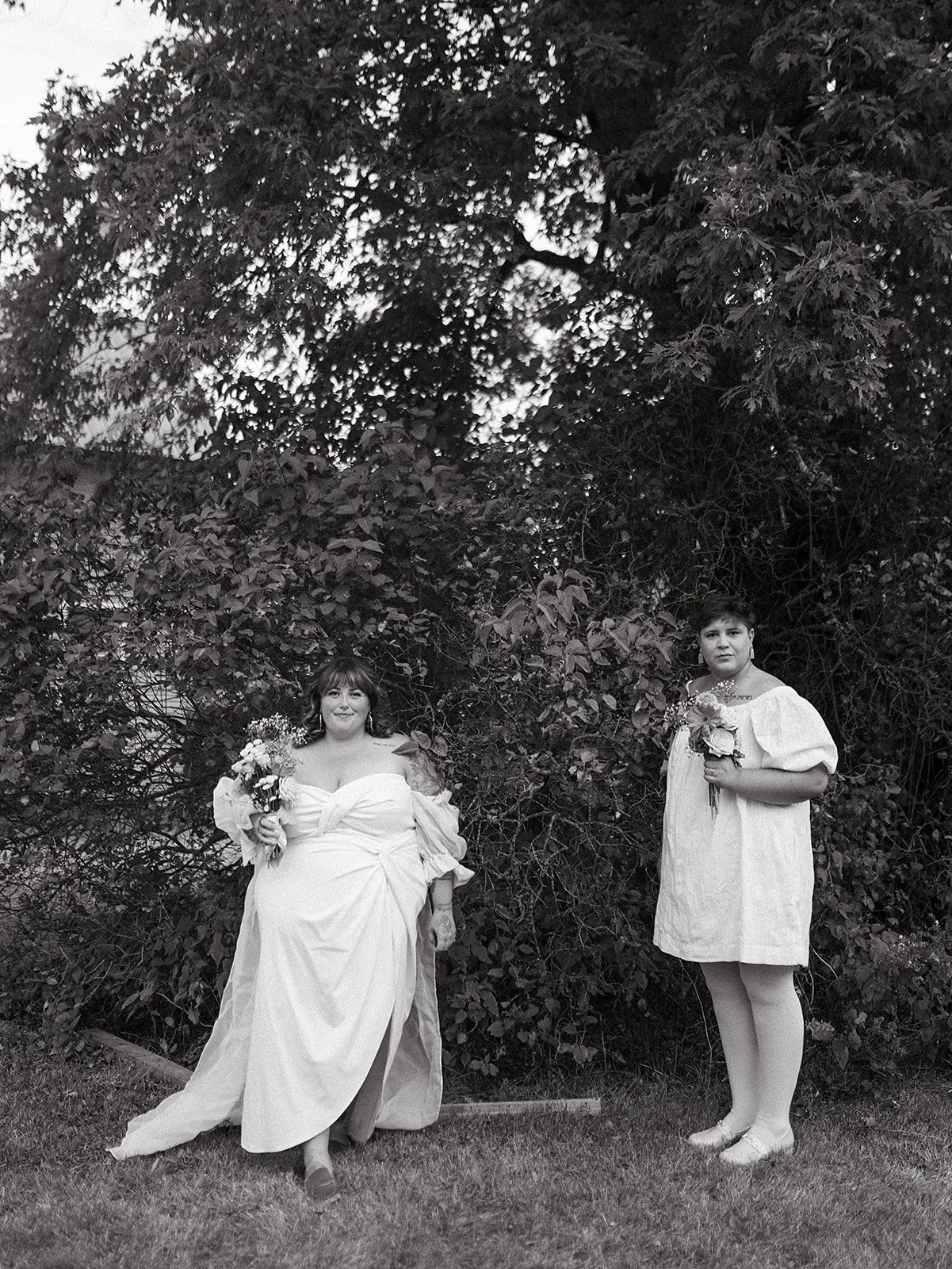 Dos mujeres con vestidos de novia sosteniendo ramos de flores, de pie frente a un arbusto grande, en un entorno exterior, en una fotografía en blanco y negro.