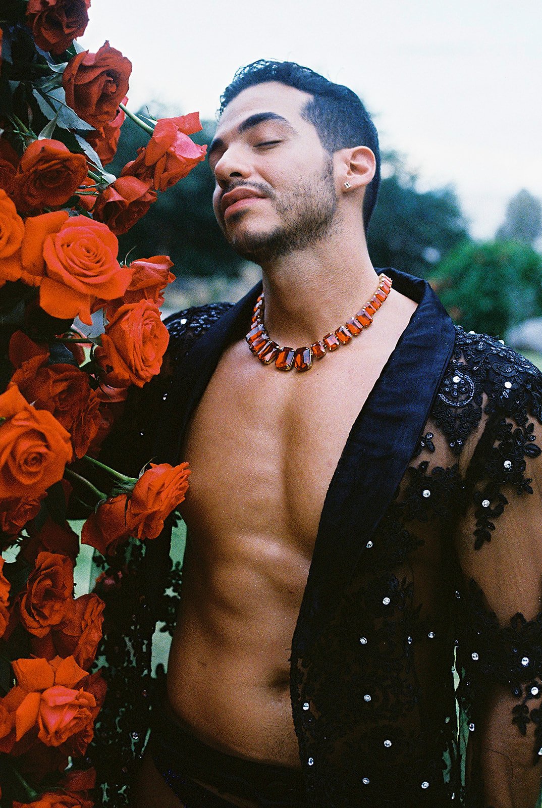 Hombre con barba, pelo oscuro y cejas pronunciadas, usando collar de piedras naranjas, vestimenta negra con detalles de encaje y pedrería, rodeado de flores naranjas y rosas. 