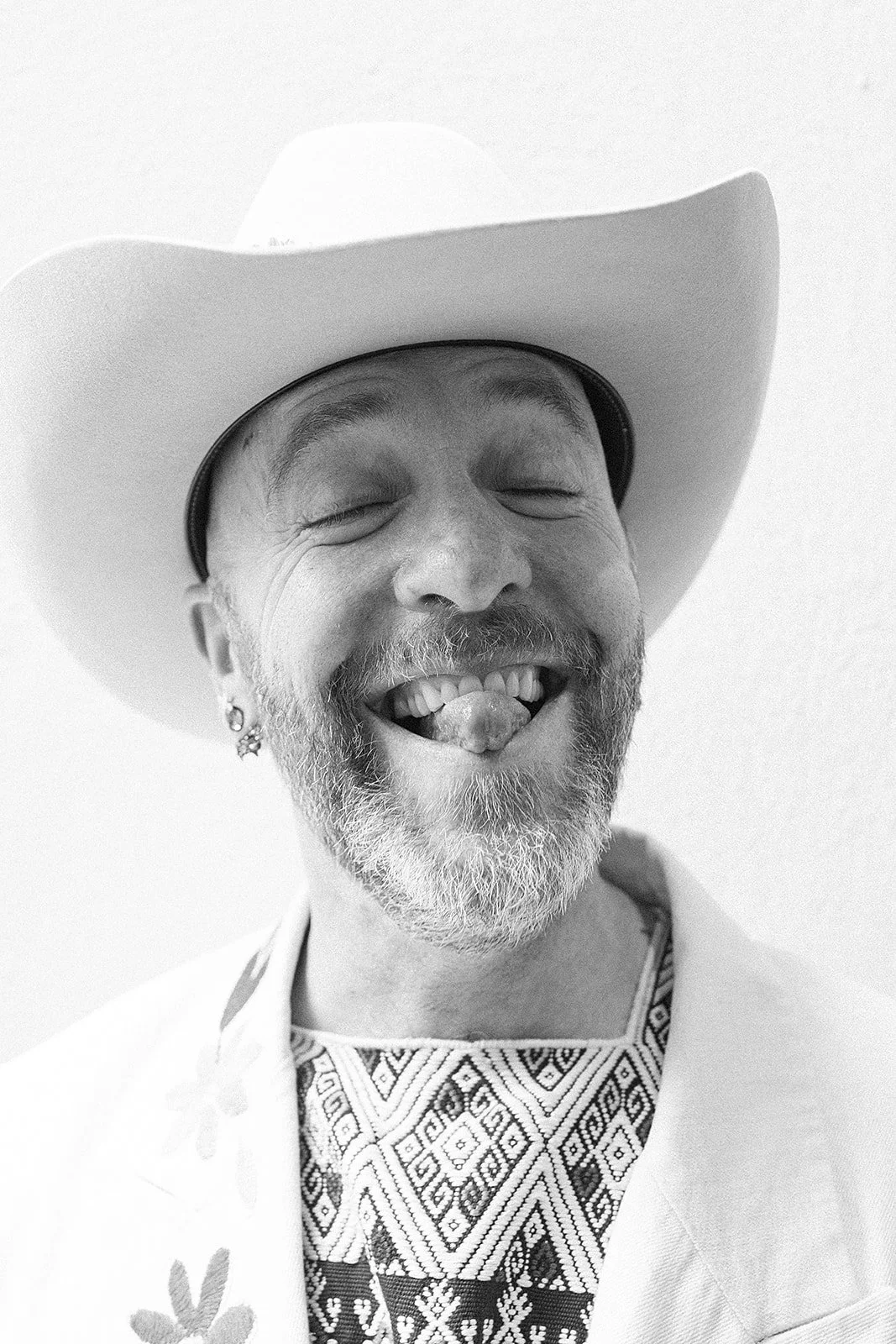 Hombre con sombrero blanco, barba gris, sonriente, con lengua afuera, usando arete en la oreja y ropa con diseño indígena en el cuello.