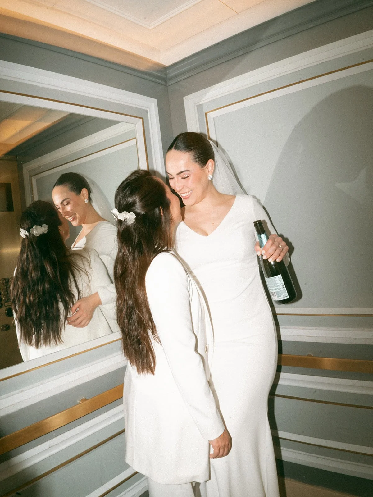 Dos mujeres vestidas de blanco son celebrando y riendo frente a un espejo, una de ellas sostiene una botella de champagne.