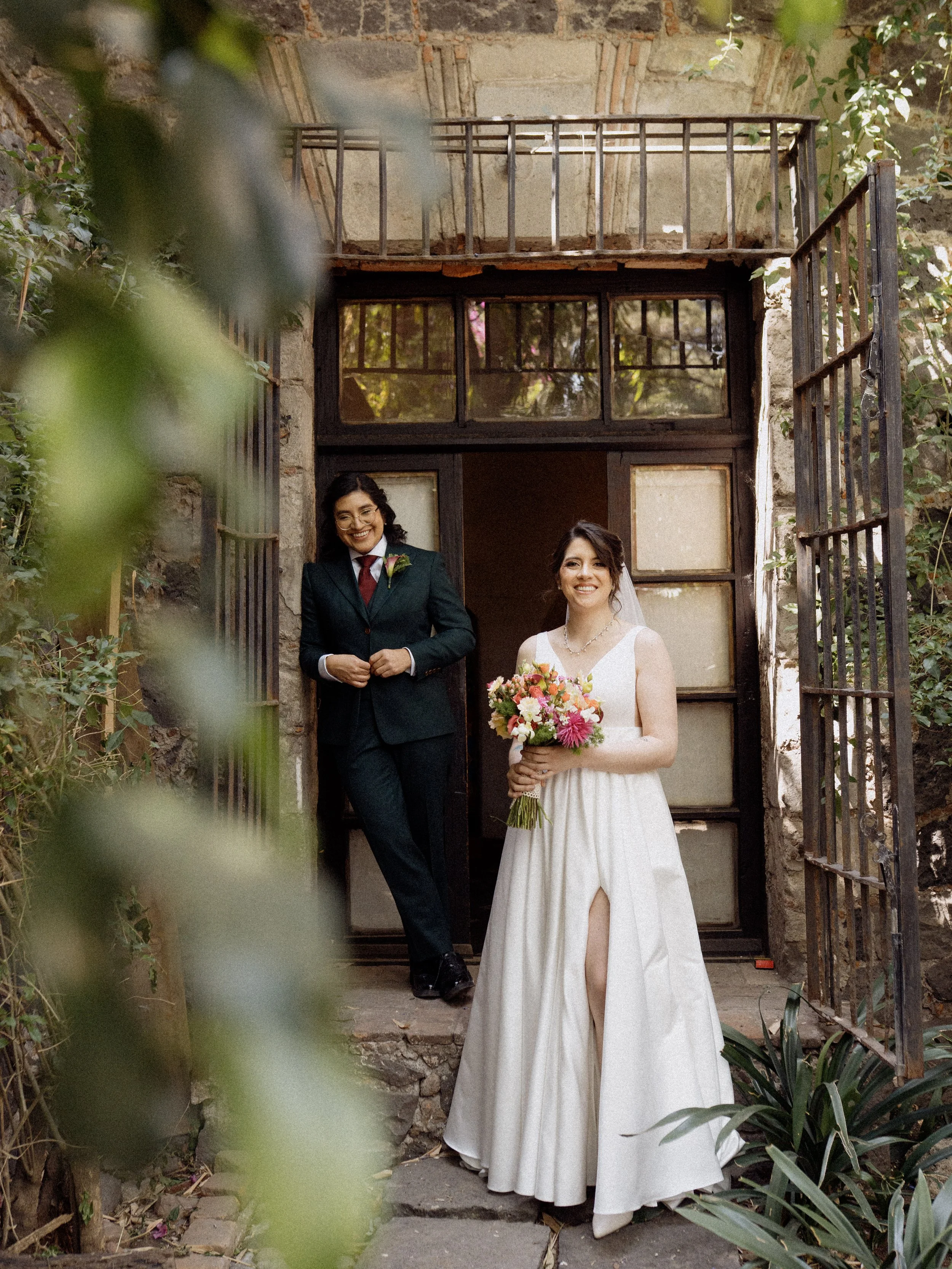Una pareja de novios posando en la entrada de una casa antigua con paredes de piedra y ventanas grandes, rodeada de vegetación.