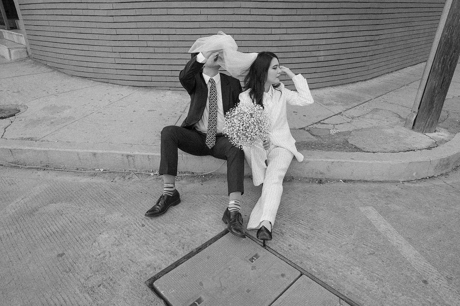Dos personas sentadas en la acera, una de ellas con traje y otra con pijama, la de traje le pone un velo a la otra, que sostiene un ramo de flores, en una calle urbana.
