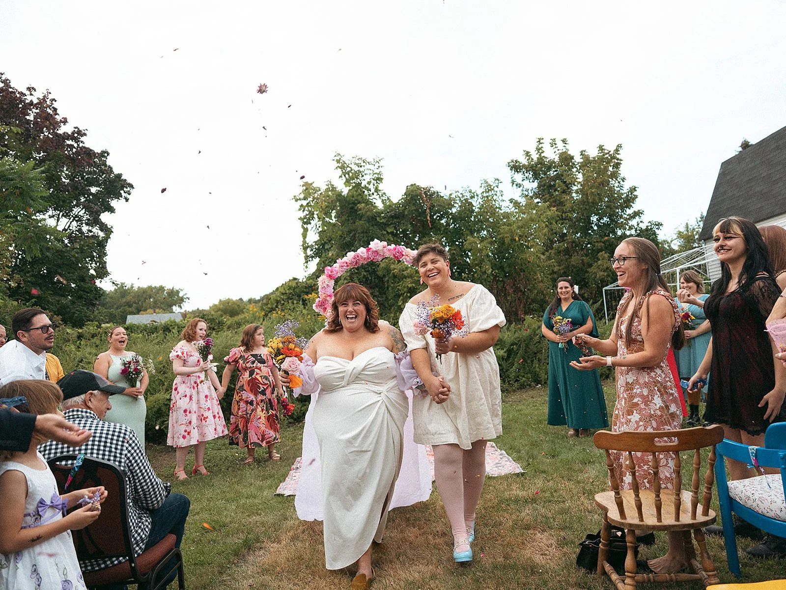 Mujeres en una boda, una de ellas vestida de blanco con flores, caminando de la mano, rodeadas de amigos y familiares en un jardín.