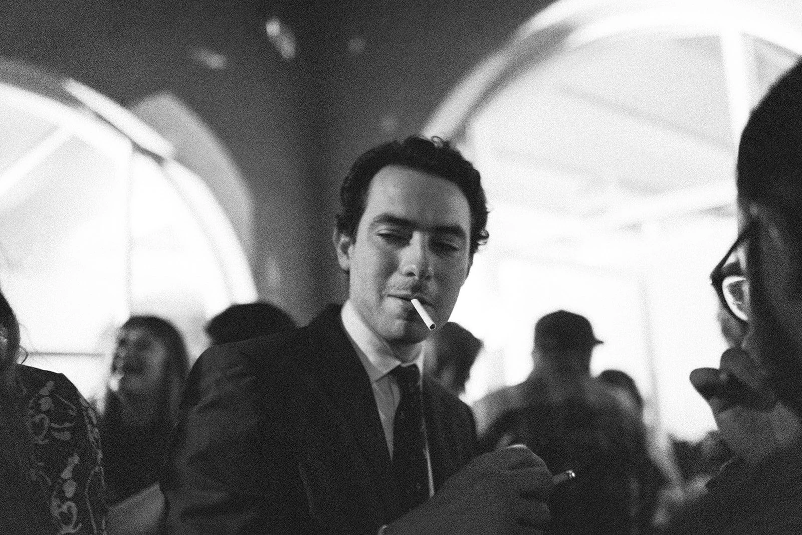 Hombre con smokings y cigarrillo en un evento social, rodeado de varias personas, en un ambiente elegante