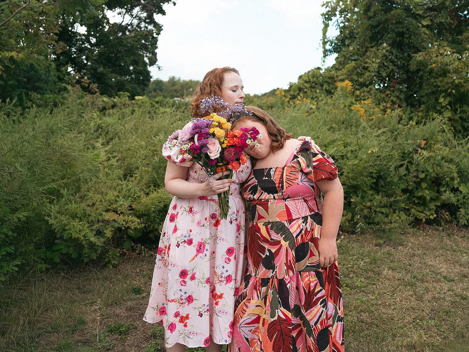 Dos mujeres con vestidos florales en un entorno natural, una de ellas con un ramo de flores y la otra con la cabeza apoyada en el ramo.