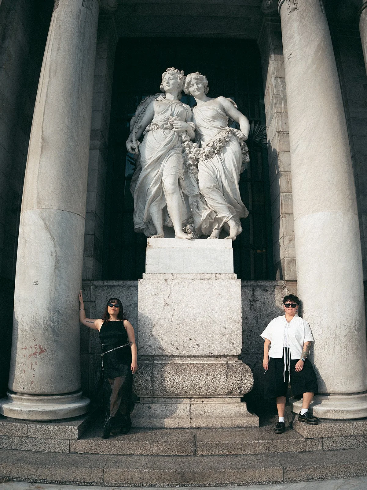 Dos personas de pie frente a una escultura clásica de mármol de dos mujeres entre columnas de piedra.