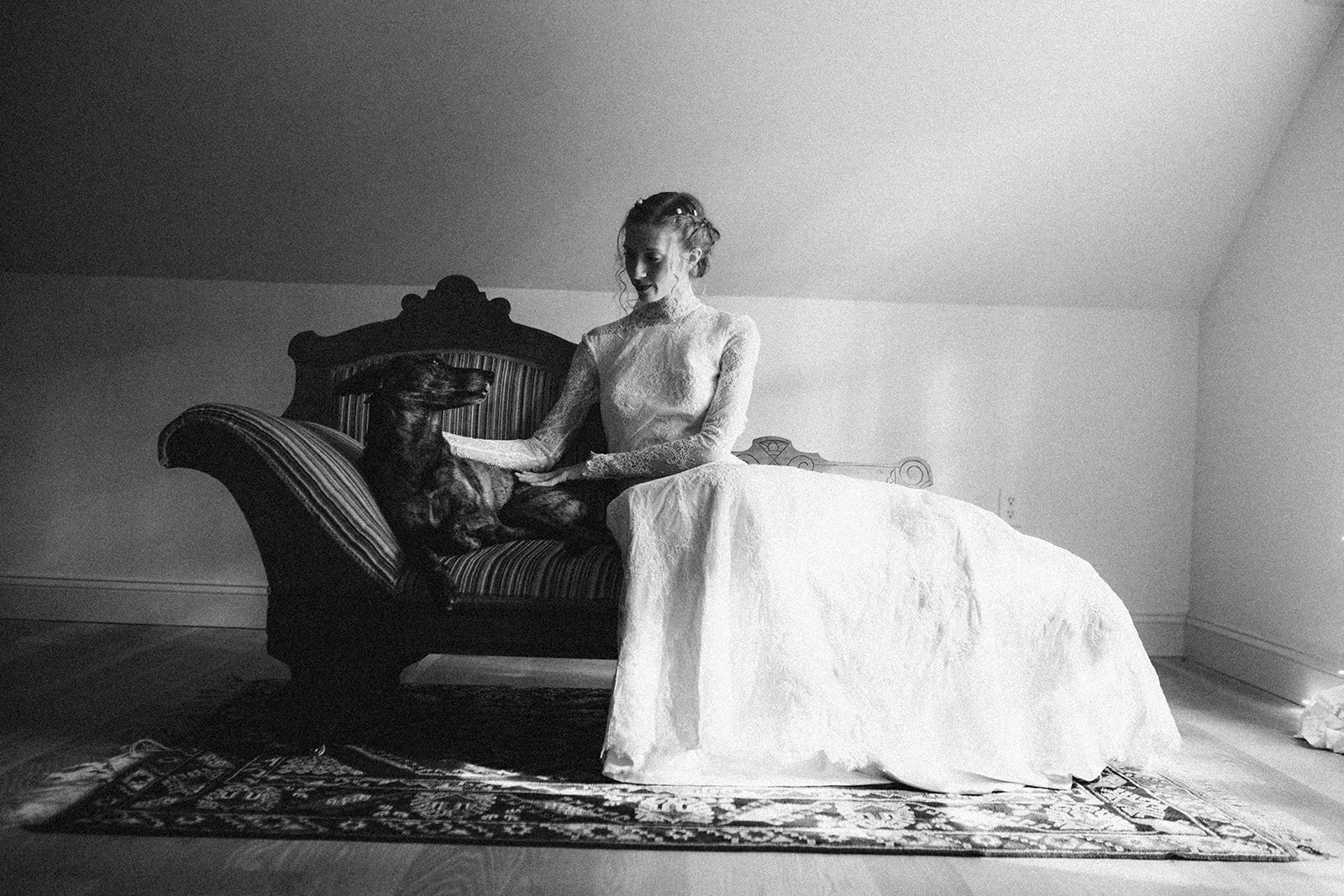 Una mujer con vestido de encaje y cabello rizado sentado en un sofá antiguo, acariciando a un perro grande acostado junto a ella en una sala sencilla, en un ambiente en blanco y negro.