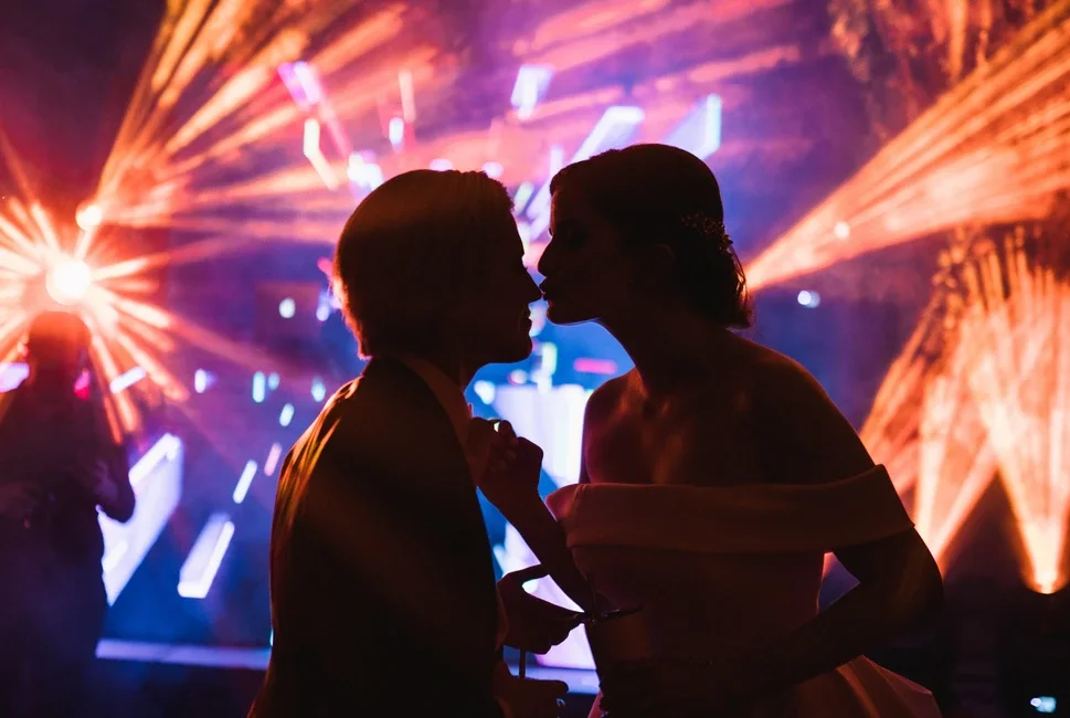 Pareja bailando y besándose en una fiesta con luces de colores y efectos de proyección en el fondo.