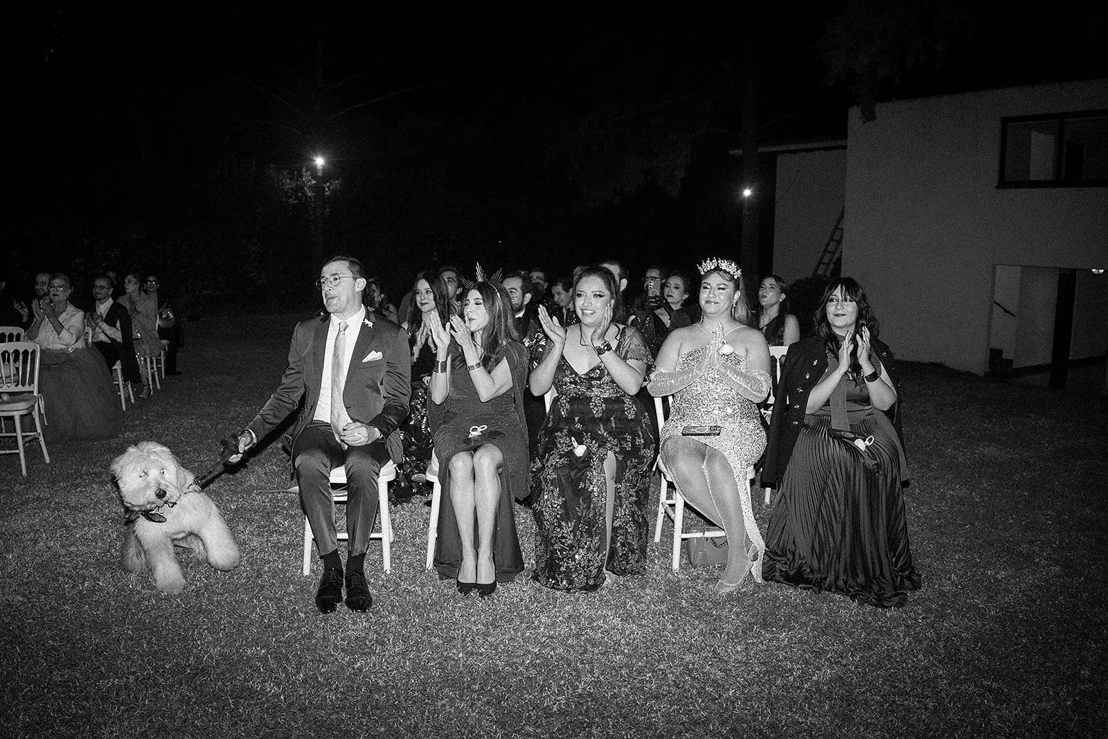 Grupo de personas sentadas en sillas al aire libre durante un evento nocturno, con varias mujeres vistiendo vestidos elegantes y una mujer con corona, y un hombre con traje y corbata, acompañados por un perro grande sentado junto a ellos.