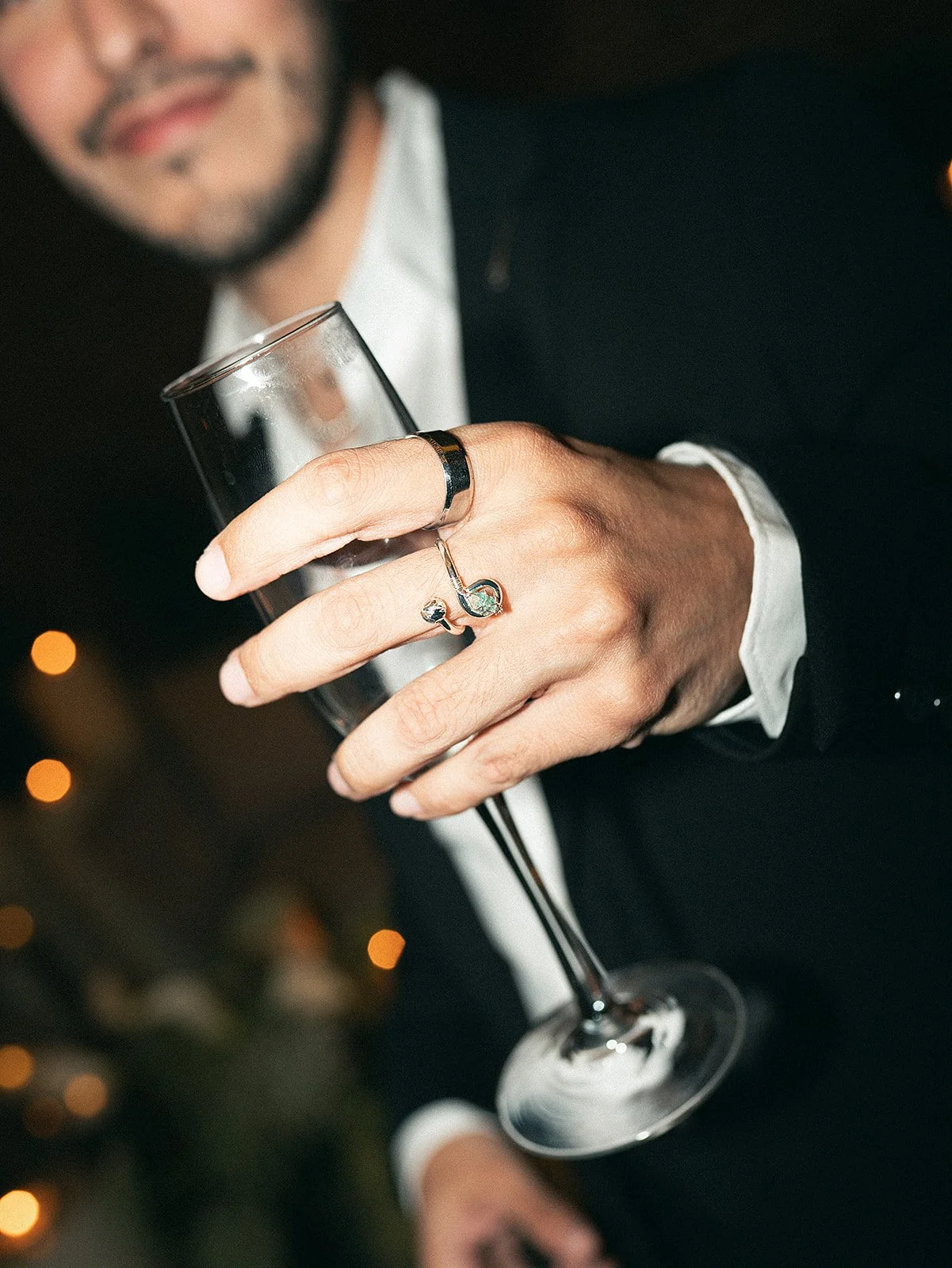 Persona con barba, vestida con ropa formal negra y camisa blanca, sosteniendo una copa de champán y usando anillos decorativos en los dedos.