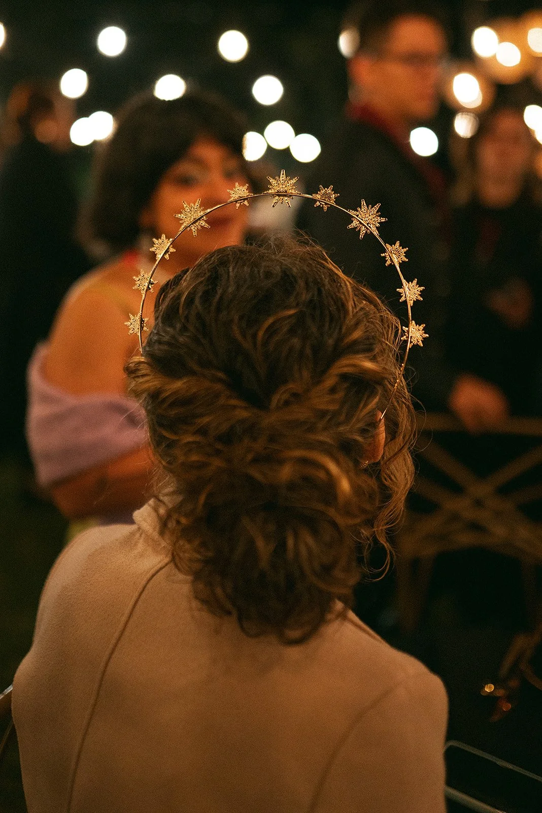 Mujer con cabello rizado y recogido, usando una tiara con copos de nieve, en un evento nocturno con luces cálidas y otras personas en el fondo.
