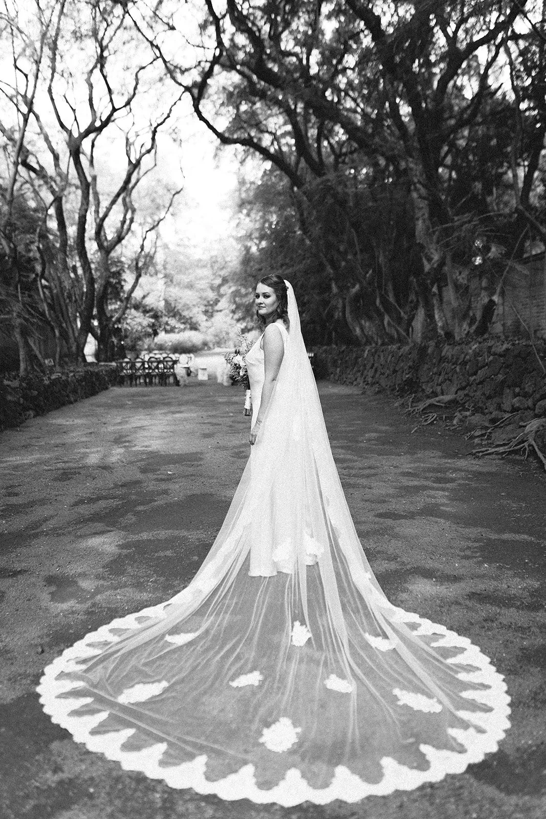 Mujer vestida de novia con vestido largo y velo, en un camino rodeado de árboles, en blanco y negro