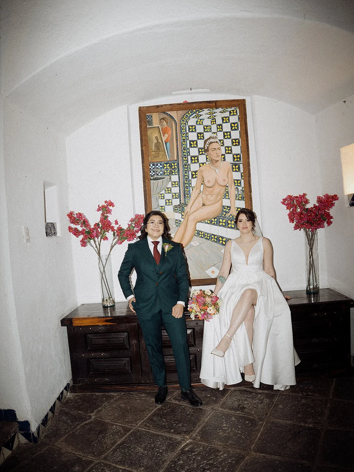 Pareja de boda, mujer con vestido blanco sentado en una mesa, hombre con traje oscuro de pie, fondo con pintura de una mujer desnuda y flores rosas en jarrones
