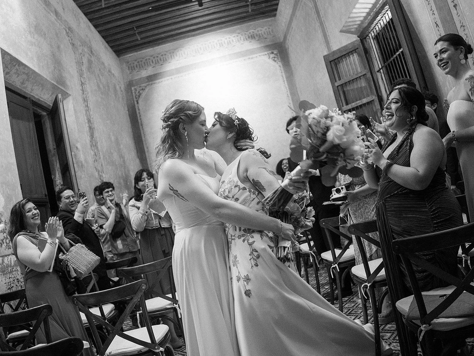 Dos mujeres en una boda besándose mientras las invitadas las observan y toman fotos, en un espacio con paredes de piedra y techo de madera.