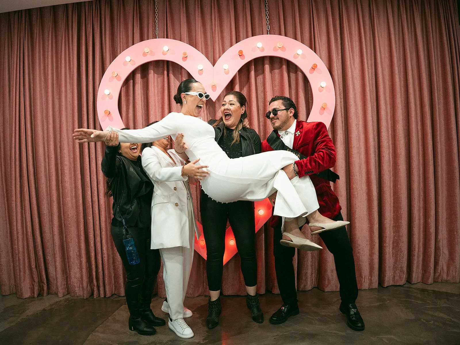 Grupo de personas en una celebración, con una mujer vestida de blanco siendo sostenida y rodeada por amigos, en un escenario con cortinas rosas y un corazón decorativo iluminado en el fondo.