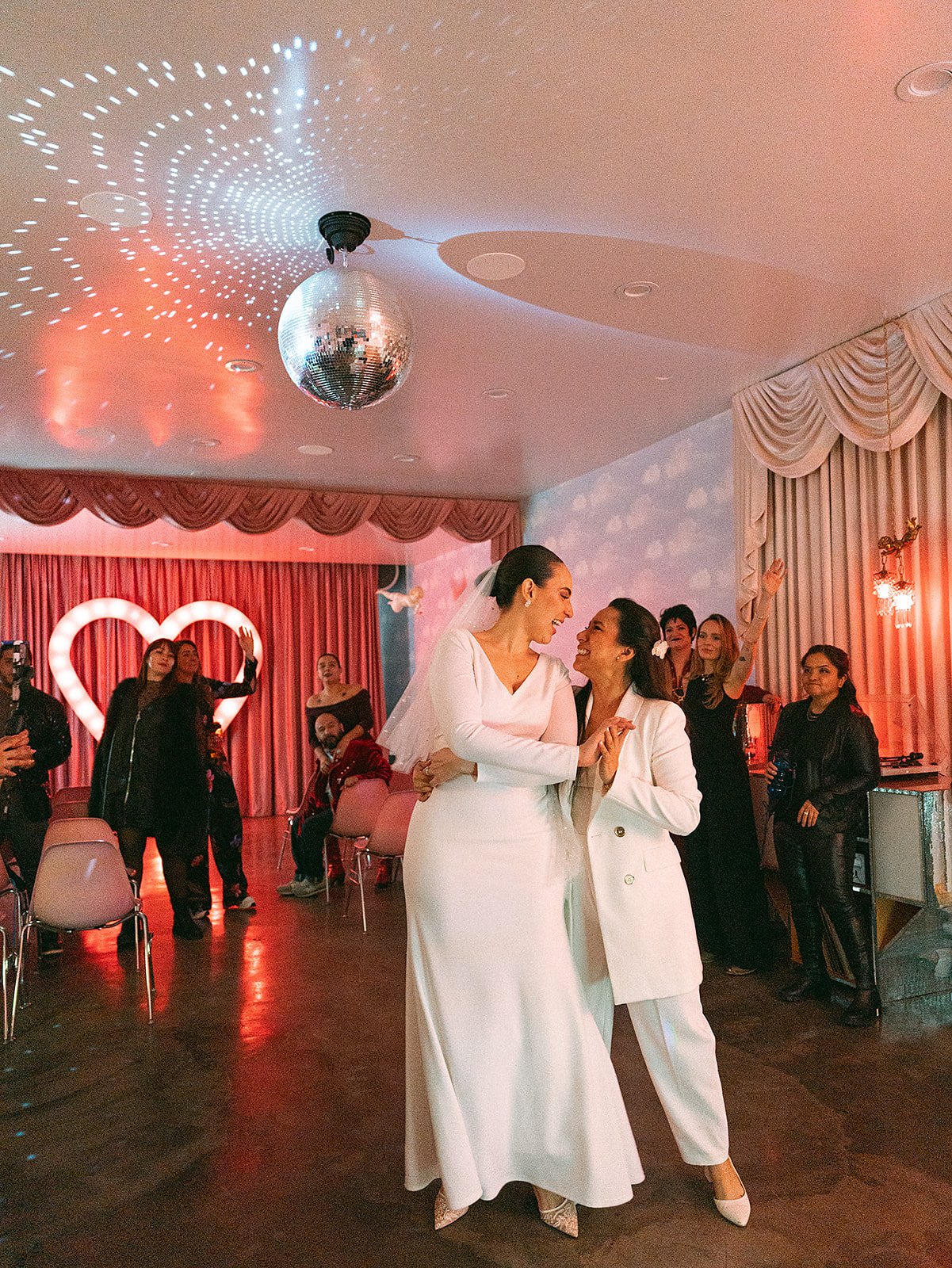 Dos mujeres en vestidos blancos bailan y se sonríen en una celebración, rodeadas de personas en un salón decorado con cortinas rosadas, luces y un corazón gigante iluminado.