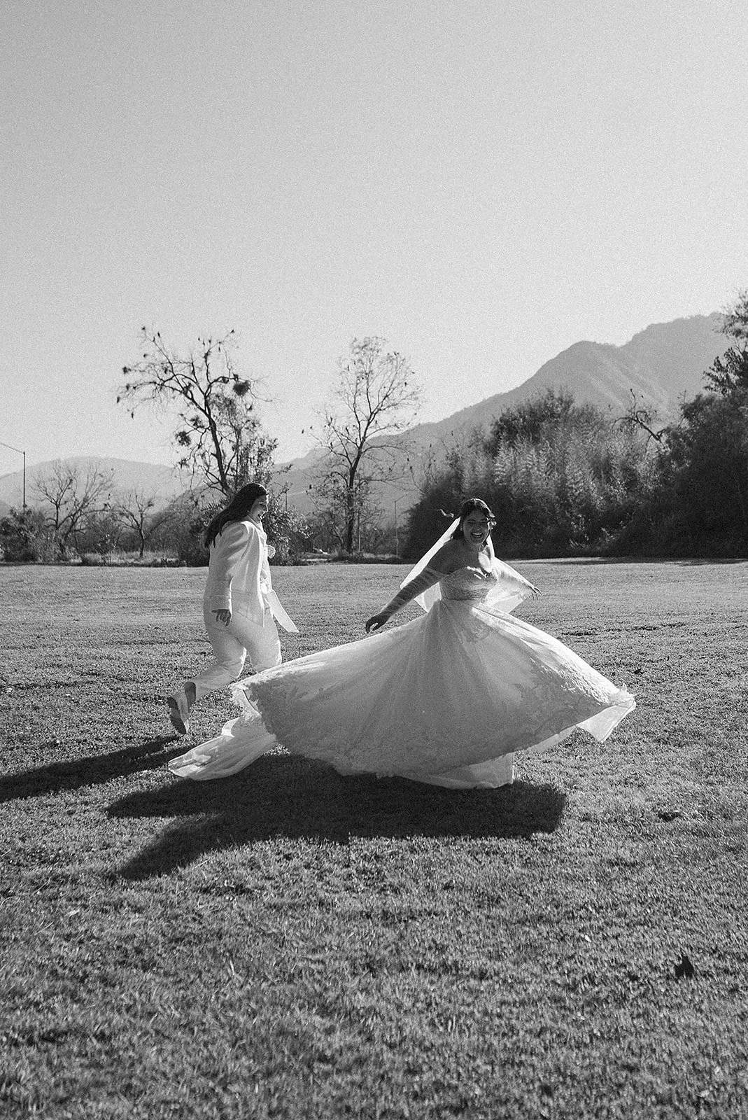 Dos personas, una en vestido de novia y otra en traje, bailando y sonriendo en un campo abierto con montañas al fondo.