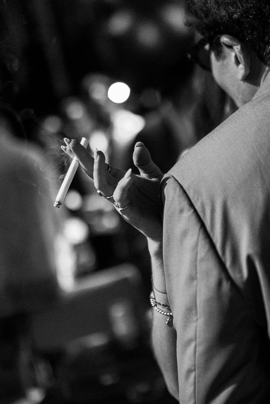 Persona sosteniendo un cigarrillo en un entorno urbano de noche, vista lateral en blanco y negro.