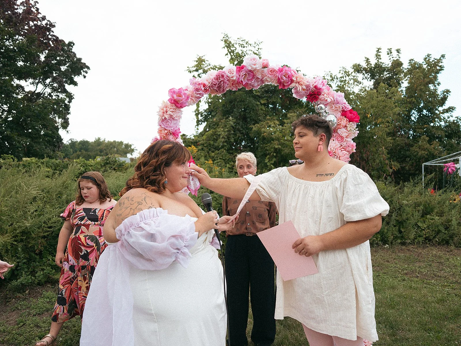 Celebración del compromiso en un entorno al aire libre con arco de flores rosas. Una pareja frente a una multitud, la mujer con cabello rizado y vestido blanco, la otra con corte de cabello corto y vestido blanco, sosteniendo un papel. Personas obser