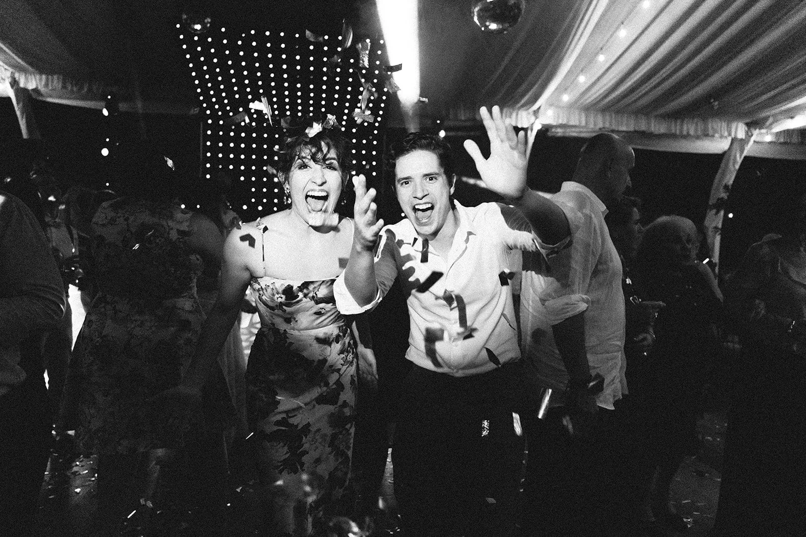 Dos personas festejando con confeti en una celebración, rodeadas de otras personas en un lugar con iluminación decorativa en el techo.