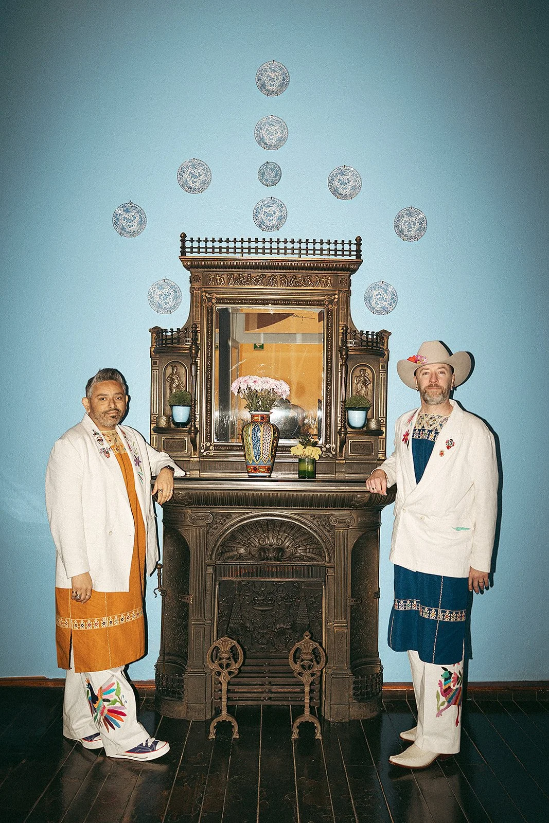 Dos hombres con ropas tradicionales mexicanas posan junto a un mueble antiguo con un espejo central y una lámpara, en una habitación con paredes color azul y decoraciones de lunares que parecen ser platos o adornos flotantes.