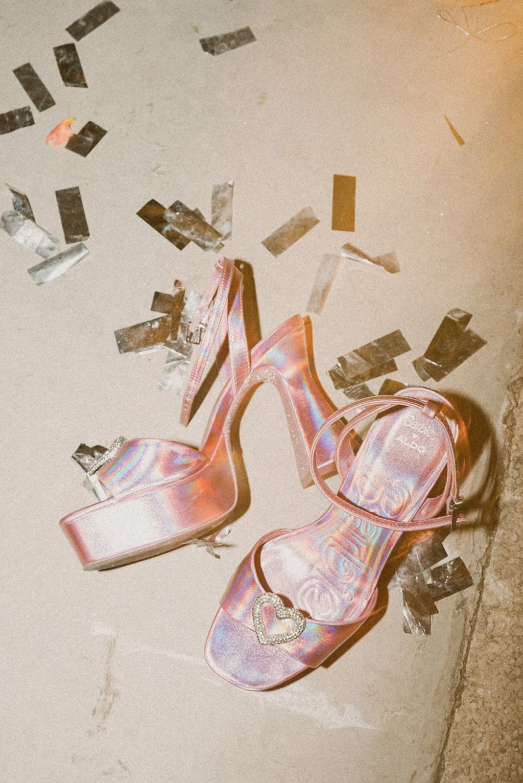 Sandalias de tacón alto de color rosa iridiscente con corazón de pedrería y una pequeña bolsa a juego, rodeadas de confeti plateado en el suelo de cemento.