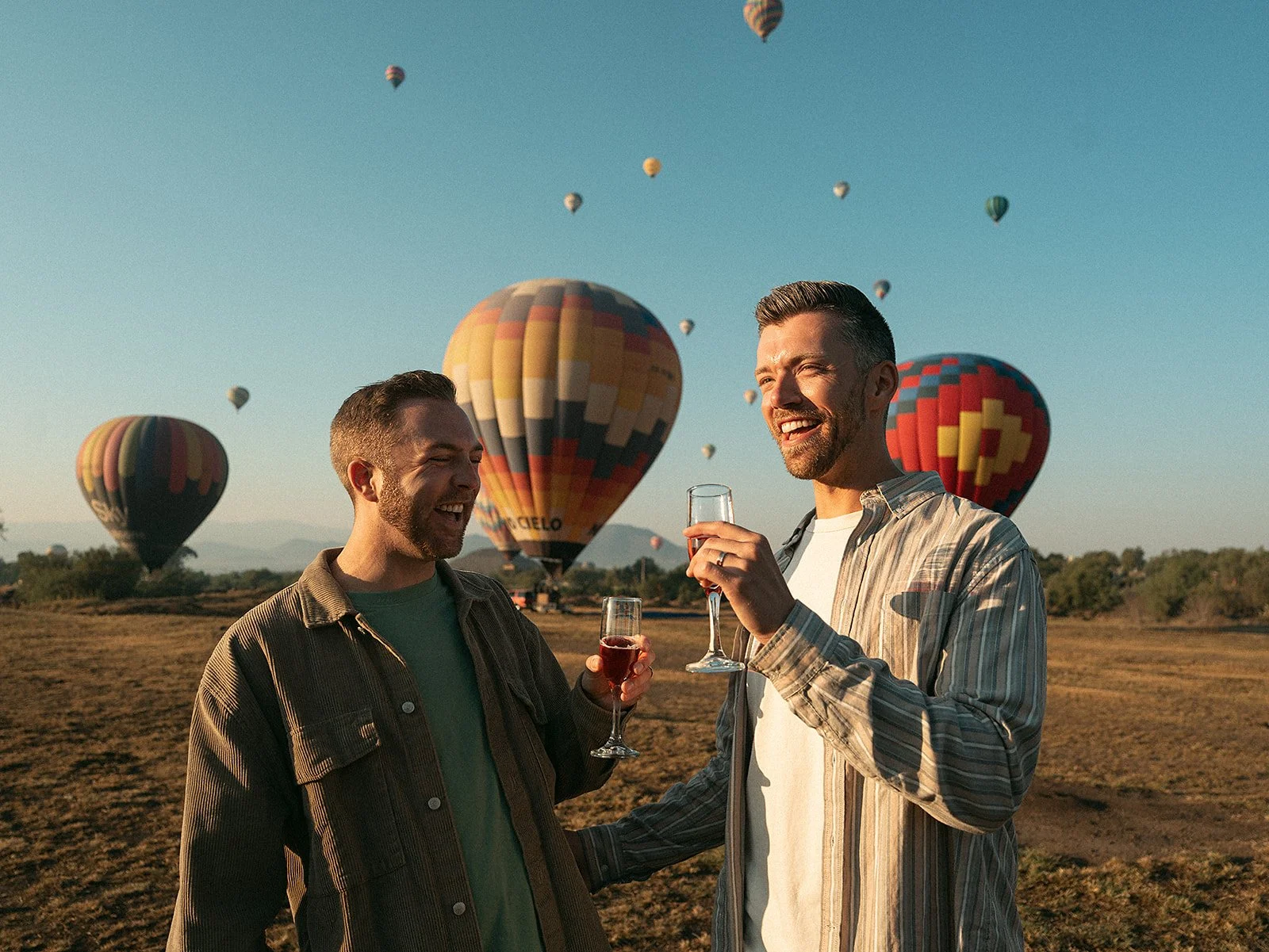 Dos hombres celebrando con copas de vino en un campo con globos aerostáticos en el cielo al atardecer.