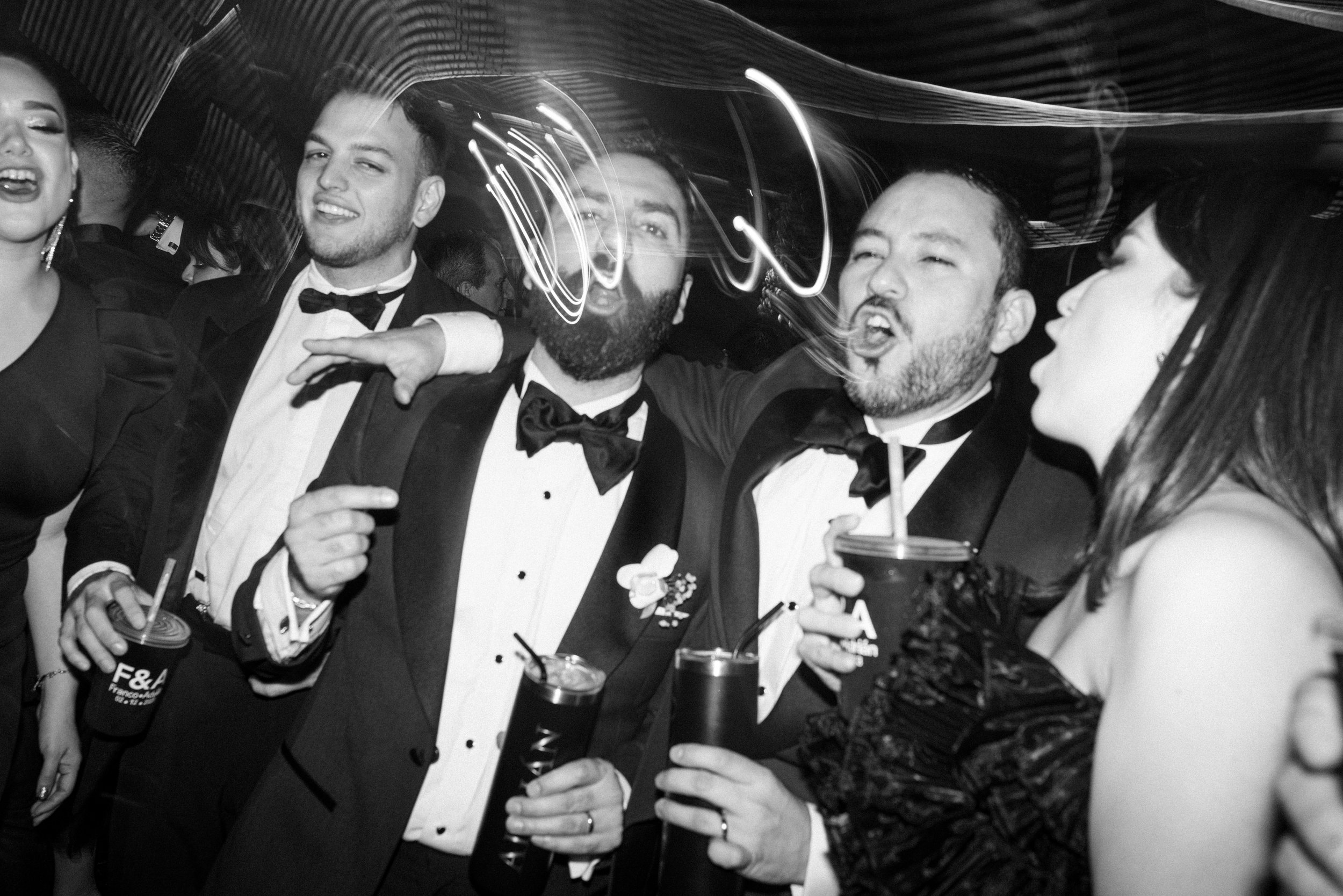 Grupo de personas en una fiesta, vestidas con trajes elegantes y tomando bebidas, disfrutando y socializando en un ambiente festivo y con luces de colores.