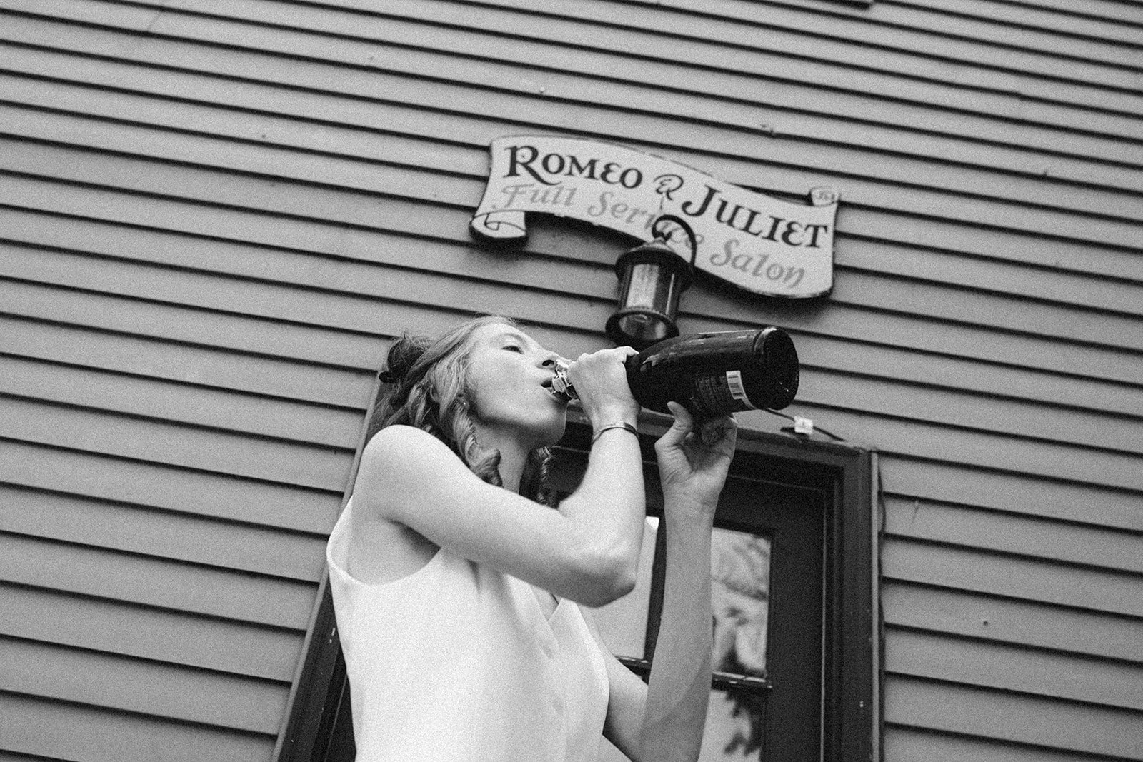 Joven mujer bebiendo una botella de vino en un lugar con pared de listones de madera y un cartel que dice "Romeo & Juliet Full Service Salon".