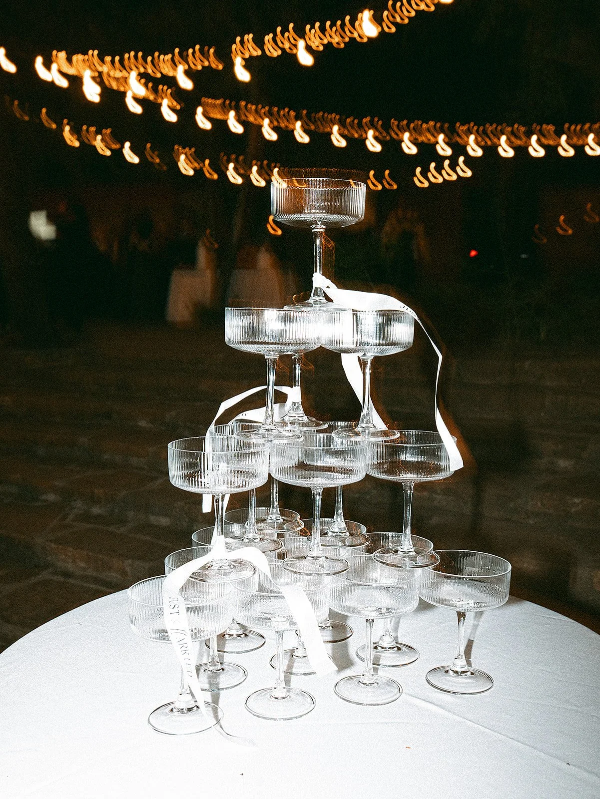 Una torre de copas de cristal apiladas en forma de pirámide en una mesa, decorada con cintas blancas, en un evento nocturno con luces decorativas en el fondo.