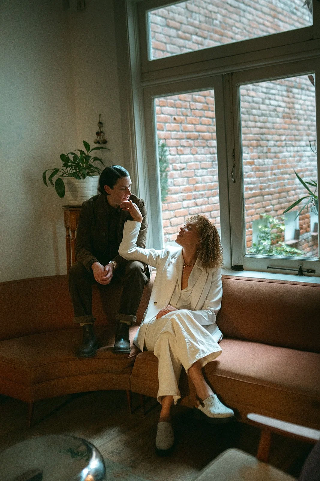 Dos mujeres en un sillón de color marrón en una habitación con gran ventana. Una mujer de cabello rizado rubio usa un traje blanco y la otra mujer de cabello oscuro con camiseta y pantalones marrones. La mujer de cabello rizado toca suavemente la car