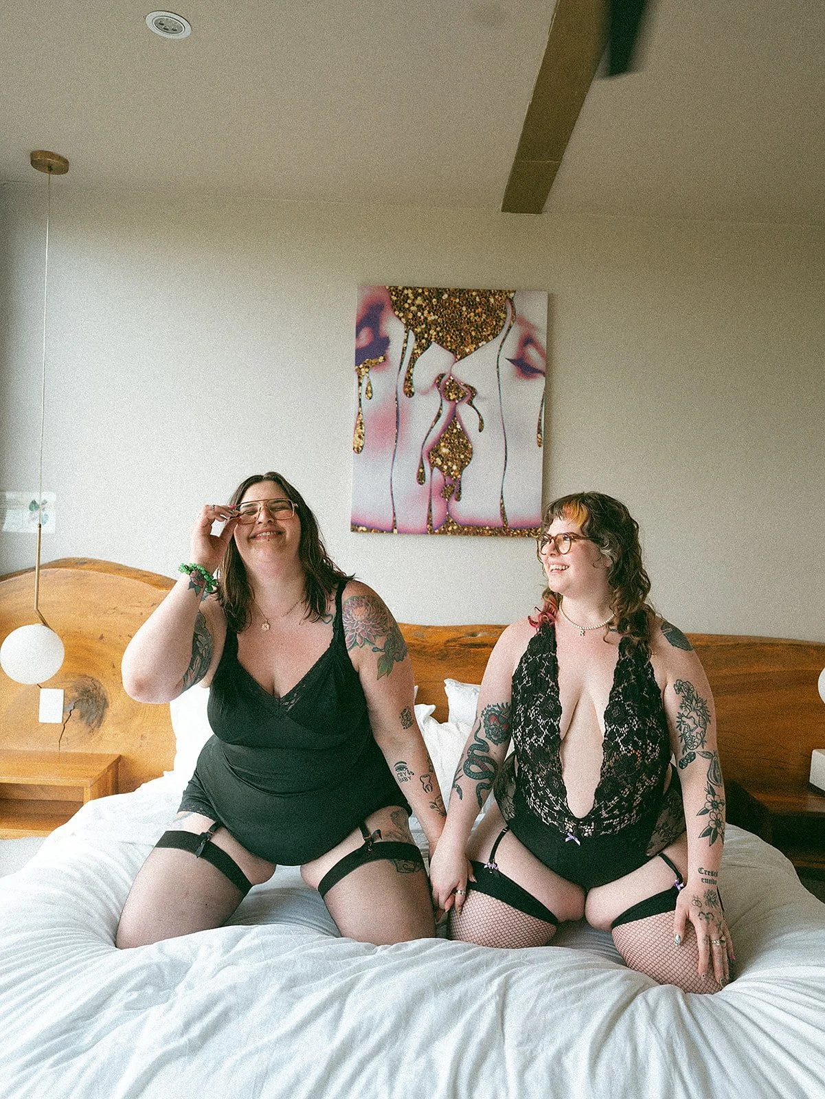 Dos mujeres en ropa de lencería negra, sentadas en una cama en una habitación moderna con arte en la pared y luces de noche.